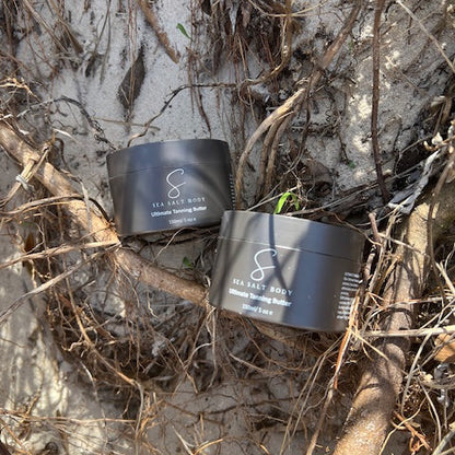 Two Sea Salt Body tanning butter jars nestled in beach driftwood and sand